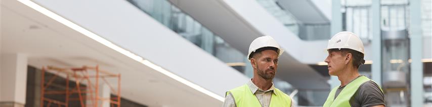 Shows two trademen discussing building works with hard hats on and high vis